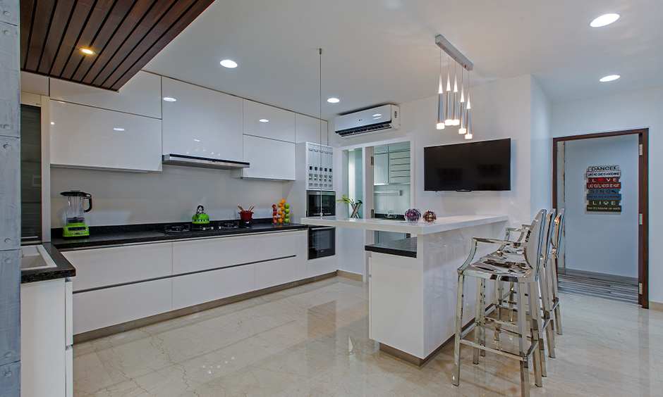 White minimalist kitchen designed in 4BHK independent house in Tumkur Road, Bengaluru with breakfast counter and steel chairs look elegant.