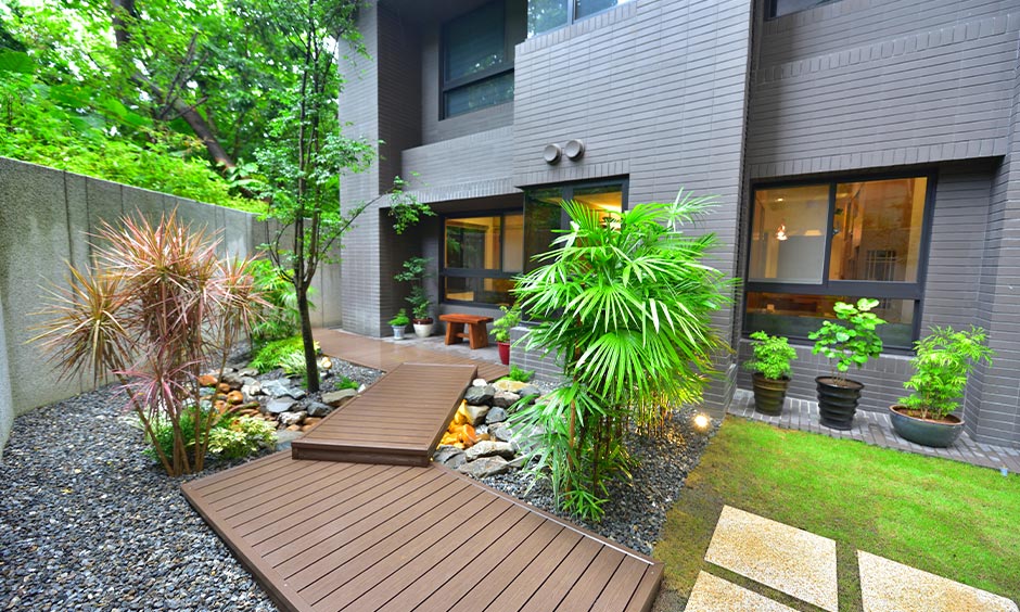 A small backyard with a wooden path and small pebbles surrounded by plants is a gardening idea for home in India.