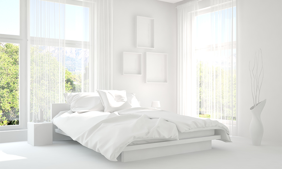 An all-white bedroom has two big windows with transparent curtains and a white vase salubrious look, white bedroom ideas.