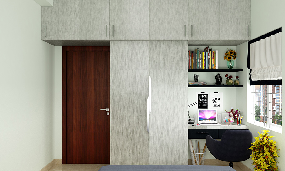 The small bedroom has a t-shaped wooden corner cupboard in grey laminate and an attached study table.