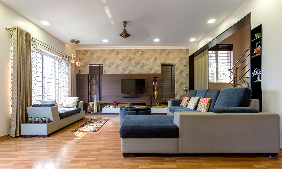 The living room in the Indian villa design has an earthy colour palette, sectional sofa, tv unit and wooden flooring.