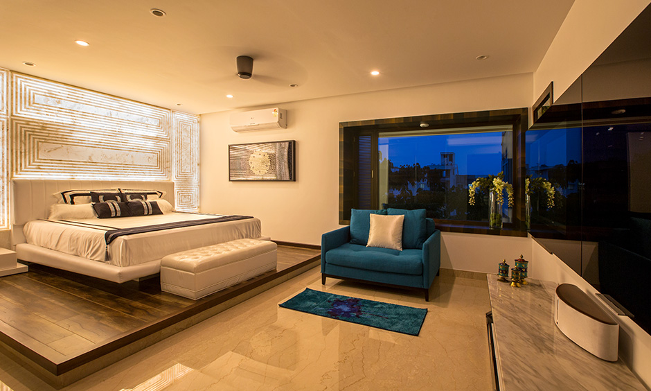 Villa bedroom in earthy colour with blue sofa and white bed with backlighting is the villa house design in India.